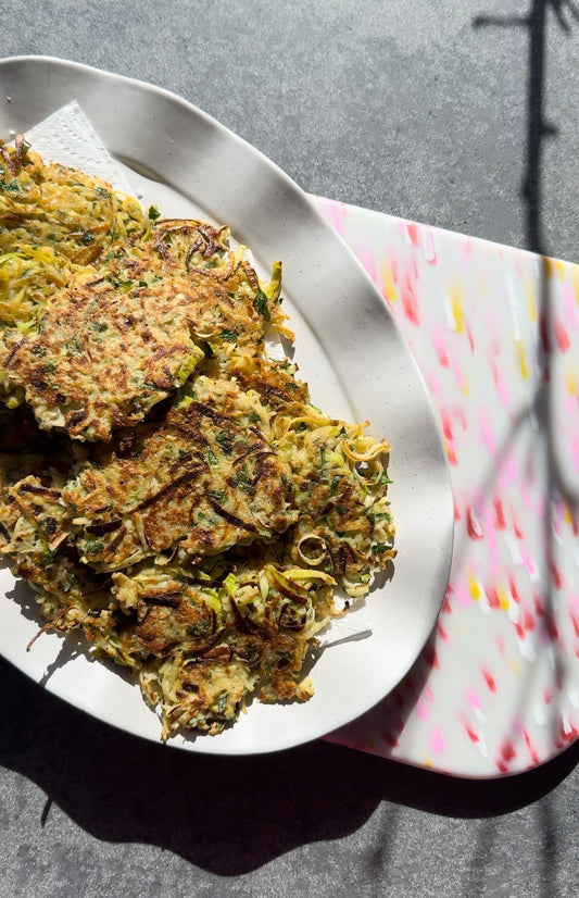 White plate with wavy edges that has 10 rosti stacked on it. the plate is sitting on a pink multicoloured chopping board