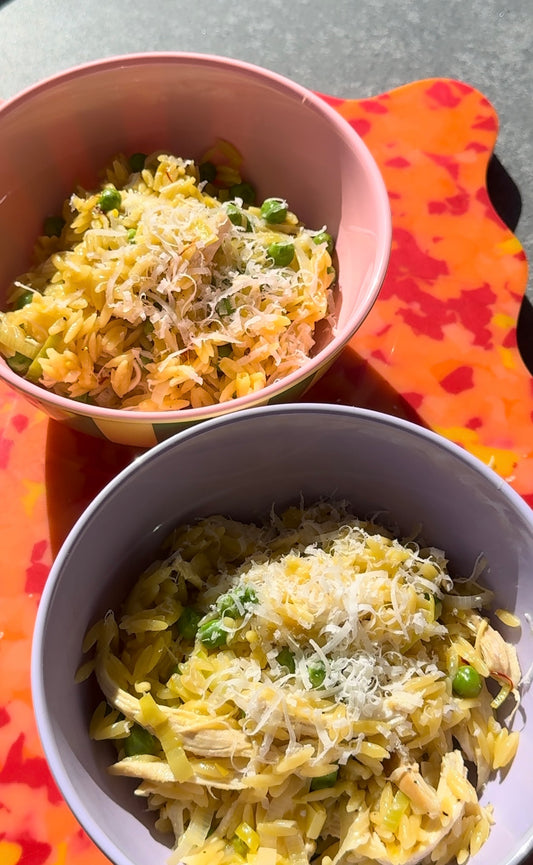 a pink and lilac bowl filled with leek, chicken and pea risoni. the bowls are sitting on a bright orange chopping board with a wavy edge.