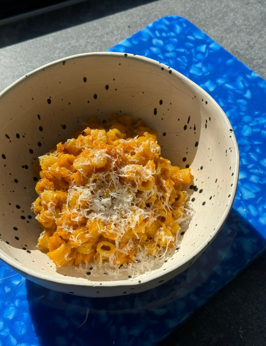 Small pasta in pumpkin sauce topped with parmesan cheese sitting inside a deep and large stone bowl with black spots. The bowl is sitting on a bright blue chopping board on a sunny benchtop.