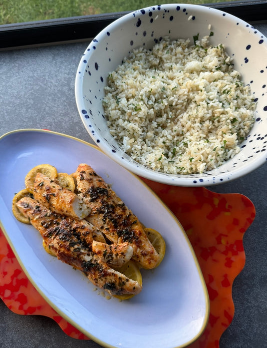 Large blue and white speckled bowl filled with garlic rice sitting next to an oval lilac plate of whiting fish that has been grilled in a lemon butter sauce sitting on a bed of lemon slices.