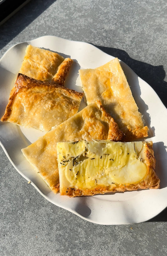 large white oval plate sitting on a grey stone bench top. The plate has 5 large slices of puff pastry loaded with sliced potatoes, rosemary and cheese.