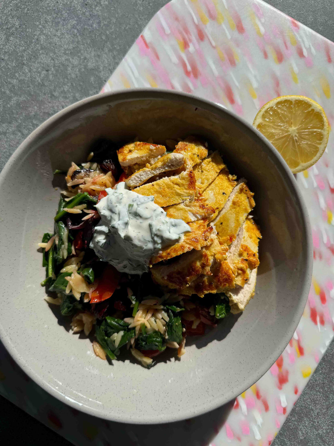 risoni pasta dish with roast veggies topped with moroccan chicken and yoghurt. Bowl of food is placed on a recylced pink and white chopping board with a lemon sliced in half