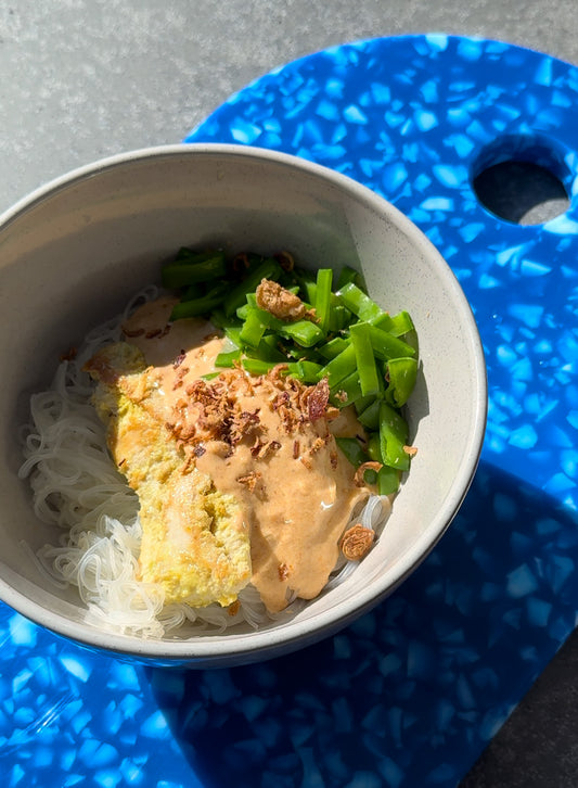 bowl of rice noodles topped with lemongrass chicken strips, satay sauce and diced snow peas. the bowl is sitting on top of a speckled blue plastic chopping board