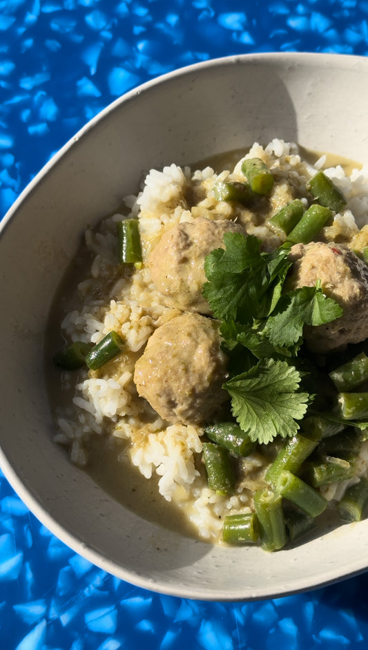 wide cream bowl filled with steamed white rice topped with Thai green curry, green beans, lemongrass meatballs and coriander. Bowl is sitting on a speckled blue chopping board.